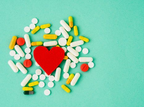 handful of pills and a red heart on a bright turquoise blue colour table, top view, copy space. Medicine help for heart health.