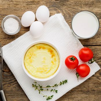 white round ramekin with oven-baked omelet of eggs and milk, with a ingredients - tomatoes, eggs, salt, glass of milk on brown rustic wooden table, top view.