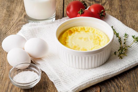 white round ramekin with oven-baked omelet of eggs and milk, with a ingredients - tomatoes, eggs, salt, glass of milk on brown rustic wooden table, side view.