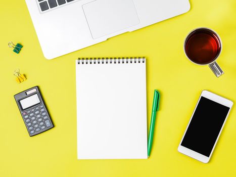 Modern creative bright yellow office desk with laptop, smartphone and other accessories. Blank Notepad page for text in the middle, top view.