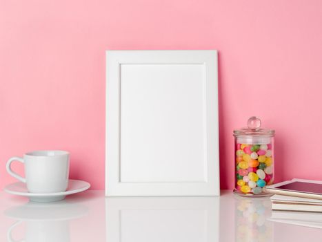 Blank white frame and candys in jar, cup of coffee or tea on a white table against the pink wall with copy space
