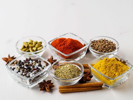 spice set-coriander, red pepper, turmeric, cinnamon, star anise, rosemary various seasonings in glass cups, on white wooden table, side view, blank space for text.