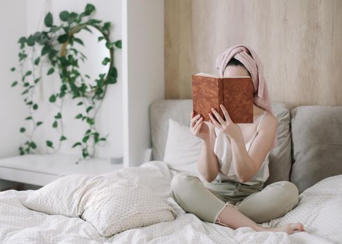 Morning book reading. Young woman with a towel on head reading book in bed in the morning. Lazy weekend at home with book. Close up, copy space, interior background.