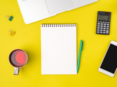 Top view of modern bright yellow office desktop with blank notepad, computer, smartphone. Mock up, empty space