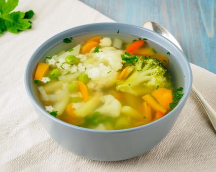 Bright spring vegetable dietary vegetarian soup with cauliflower, broccoli, pepper, carrot, green peas, pasta, parsley. Side view, blue background.