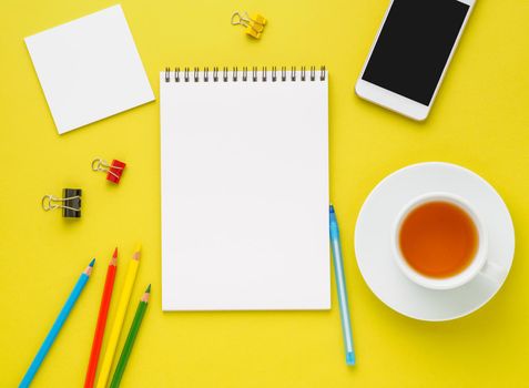 Top view of modern bright yellow office desktop with blank notepad, cup of tea and supplies. Mock up, empty space