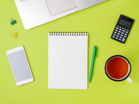 Modern creative bright green office desk with laptop, smartphone and other accessories. Blank Notepad page for text in the middle, top view.