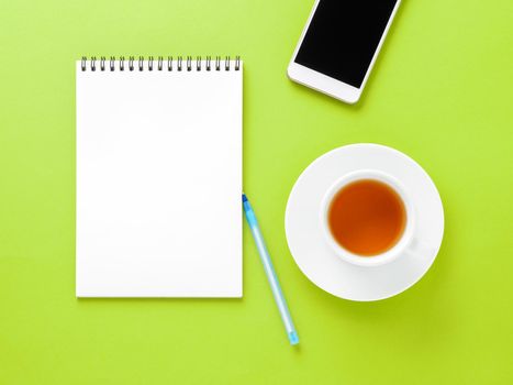 Top view of modern bright green office desktop with blank notepad, cup of tea, smartphone. Mock up, empty space