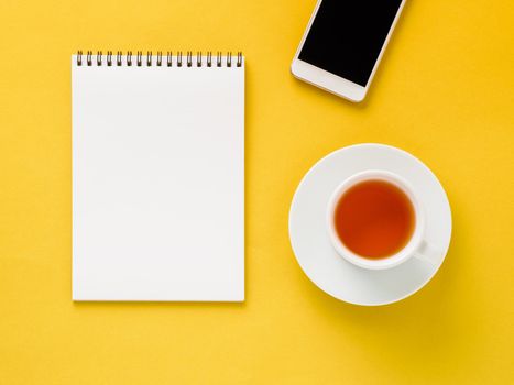 Top view of modern bright yellow office desktop with blank notepad, cup of tea, smartphone. Mock up, empty space