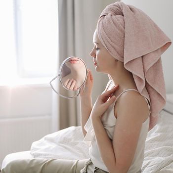 young woman with a towel on head looking in the mirror in a cozy sunny room at home.
