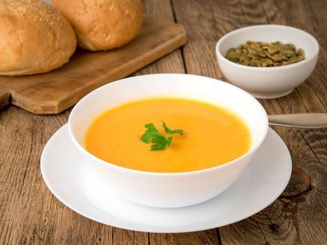 white bowl of pumpkin soup, garnished with parsley on wooden background, side view.