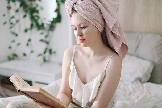 Morning book reading. Young woman with a towel on head reading book in bed in the morning. Lazy weekend at home with book. Close up, copy space, interior background.