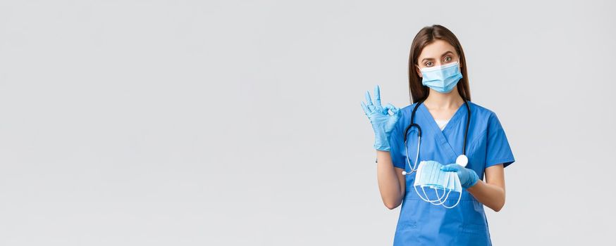 Covid-19, preventing virus, health, healthcare workers concept. Professional female nurse or doctor in blue scrubs and personal protective equipment, show okay sign and give medical mask.