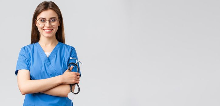 Healthcare workers, medicine, insurance and covid-19 pandemic concept. Confident and enthusiastic young female nurse, doctor in blue scrubs and glasses, hold stethoscope, smiling camera pleased.