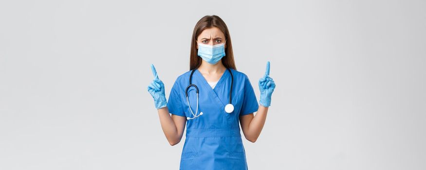 Covid-19, preventing virus, health, healthcare workers and quarantine concept. Skeptical and disappointed female nurse in blue scrubs feel displeased, pointing fingers up, wear medical mask.