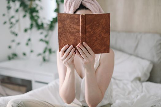 Morning book reading. Young woman with a towel on head reading book in bed in the morning. Lazy weekend at home with book. Close up, copy space, interior background.