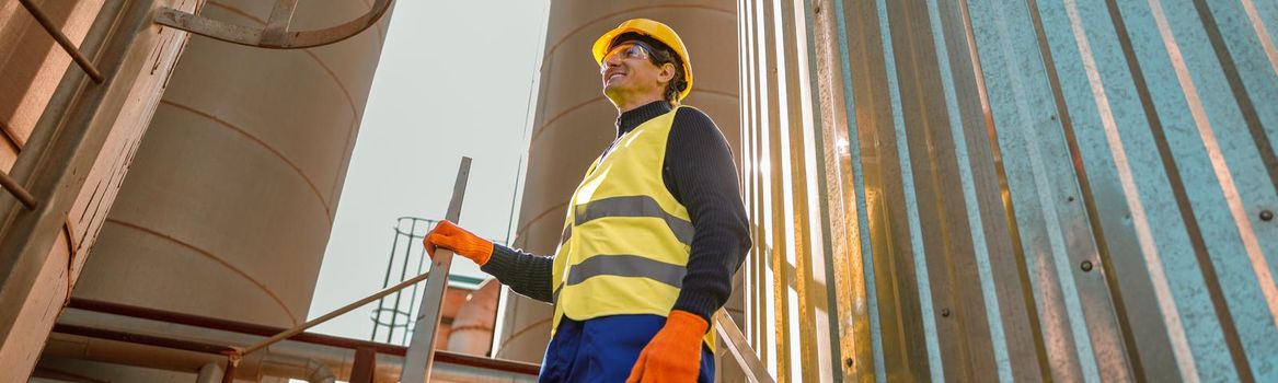 Smiling matured man engineer wearing safety helmet and work vest while standing among metal storage containers at industrial plant