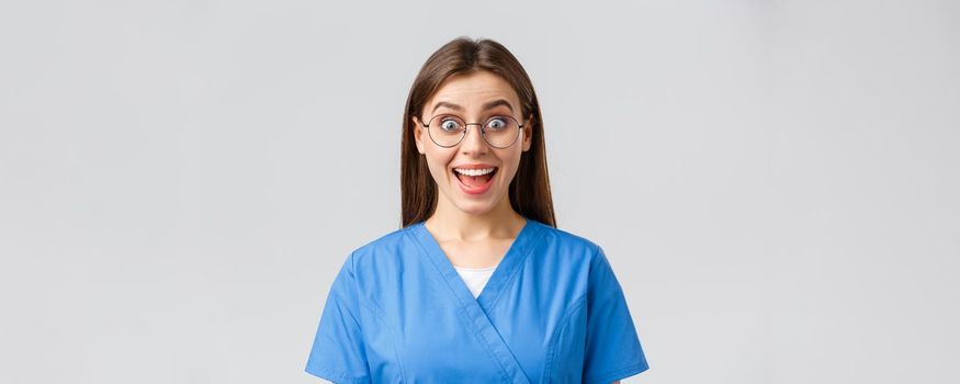 Healthcare workers, medicine, insurance and covid-19 pandemic concept. Enthusiastic upbeat female nurse, doctor in blue scrubs and glasses hear fantastic news, smiling amused.