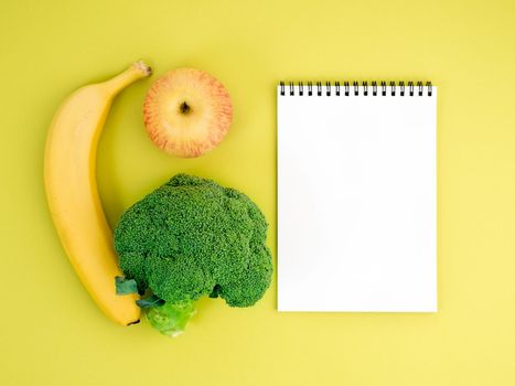 Fruits and vegetables - apple, banana and broccoli on bright yellow background. Notebook to record about proper nutrition, vitamins, healthy lifestyle.