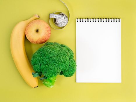 Fruits and vegetables - apple, banana and broccoli on bright yellow background. Notebook to record about diet.