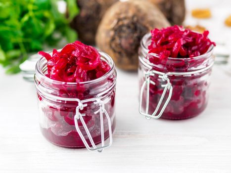 Fresh salad of grated boiled beetroot in glass jars, white wooden background, side view.