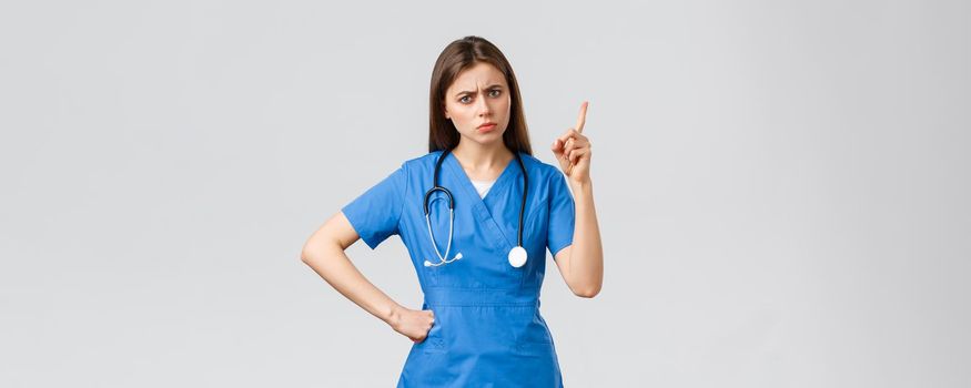 Medical workers, healthcare, covid-19 and vaccination concept. Frowning young female nurse or doctor in blue scrubs, frowning, shaking finger as scolding patient bad behaviour, grey background.