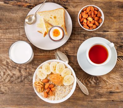 Breakfast in the morning, on a wooden table, top view. A glass of milk, tea, oatmeal with fruits and nuts, soft-boiled egg.