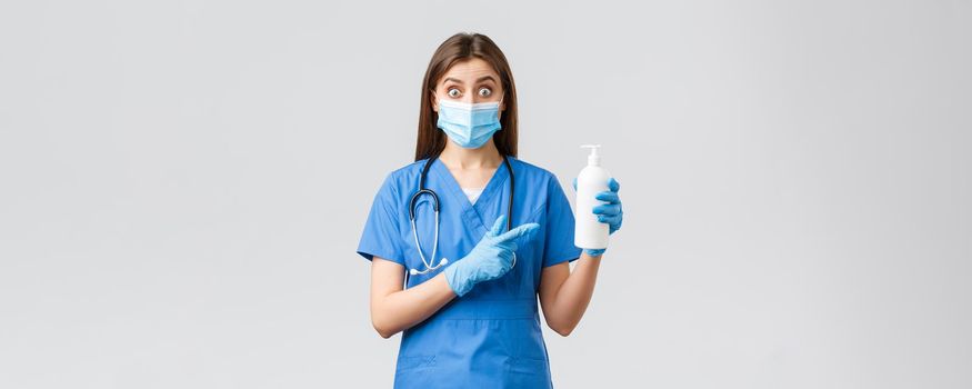 Covid-19, preventing virus, health, healthcare workers and quarantine concept. Surprised female nurse, cute doctor in blue scrubs, medical mask and gloves pointing finger at hand sanitizer or soap.