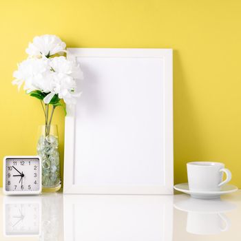 White frame, flower in vase, cup with tea or coffee, clock on a white table against yellow wall with copy space. Mock up.