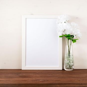 White frame, flower in glass bottle on a dark brown wooden table against the white wall with copy space. Mock up.