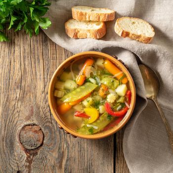 Bright spring vegetable dietary vegetarian soup with potatoes, pepper, carrot, green peas, parsley. Top view, brown rustic wood background, linen napkin.