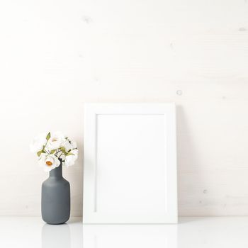 Blank white frame, flower in vaze on a white table against the white wall with copy space. Mock up.