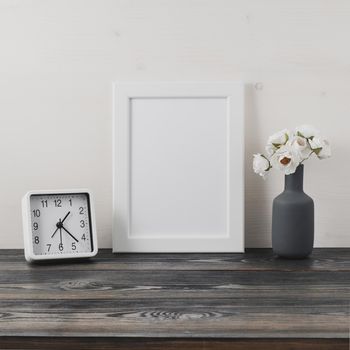 White frame, flower in vaze, clock on a dark gray wooden table against the white wall with copy space. Mock up.