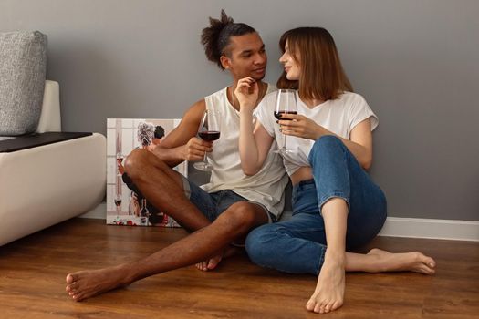 Stylish, modern, multiracial couple, a guy and a girl in white t-shirts and blue jeans, sit at home on the floor against a gray wall, hold glasses of red wine in their hands, hug. copy space