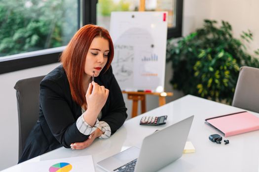 Young red-haired girl teleworking from home in her living room with her white laptop. Brightly lit scene with a large window in the background with natural light. She is wearing a black blazer. Young self-employed businesswoman and entrepreneur working at computer.