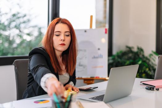 Pretty young redheaded pretty woman teleworking from home. She is making a work video call explaining something. Very bright environment with a large window behind and natural light. Young businesswoman and entrepreneur working on white laptop.