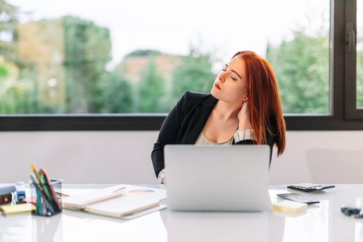 Young red-haired girl in grey T-shirt and black blazer teleworking from home. Enterprising and self-employed businesswoman working in the office or at home. She is stretching her back due to many hours of work at the computer. Large window in background with natural light.