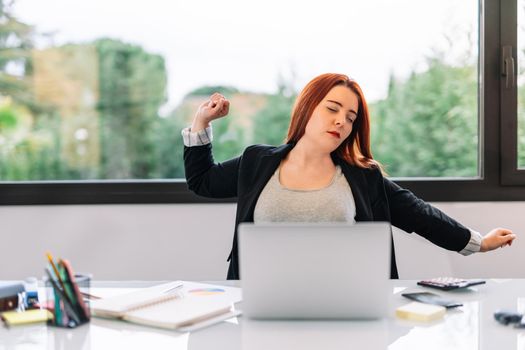 Young red-haired girl in grey T-shirt and black blazer teleworking from home. Enterprising and self-employed businesswoman working in the office or at home. She is stretching her back due to many hours of work at the computer. Large window in background with natural light.