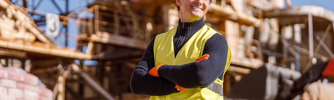 Joyful male engineer wearing safety helmet and work vest while keeping arms crossed and smiling