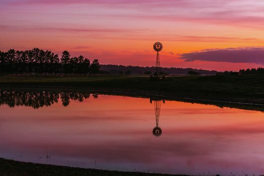 Old farm windmill in a rural field with vivid sunrise sky of reds and orange colours.