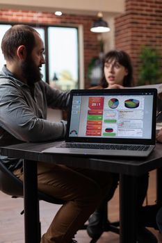 Laptop computer having marketing project graphs on screen while multiethnic businesspeople discussing company turnover in startup office. Diverse team planning business collaboration