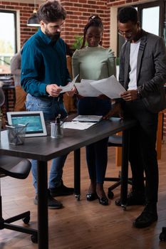 Diverse team holding papers analyzing marketing graph discussing business project in brick wall office. Multi-ethnic businesspeople brainstorming ideas working at company strategy. Corporate staff