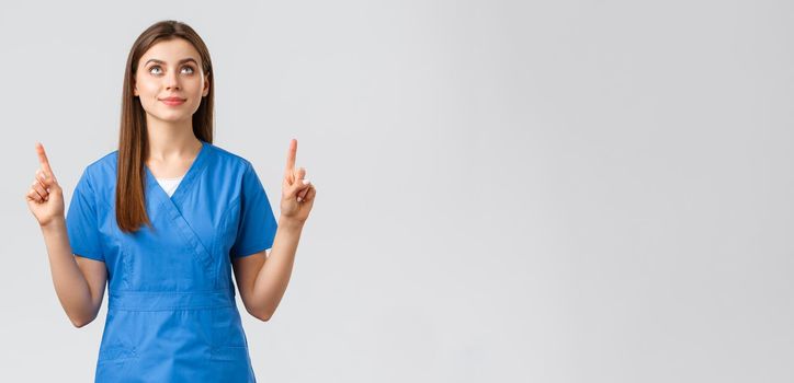 Healthcare workers, prevent virus, covid-19 test screening, medicine concept. Young talented doctor, female nurse in blue scrubs, pointing fingers up and looking at sign or banner, grey background.