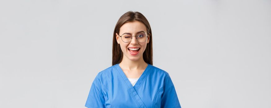 Healthcare workers, medicine, insurance and covid-19 pandemic concept. Cheeky pretty female nurse, intern or doctor in clinic, wear scrubs and glasses, wink at camera with enthusiastic smile.