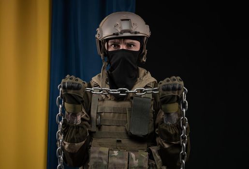 a male soldier in a military uniform and helmet with a weapon on the background of the national flag of Ukraine with a chain as a symbol of slavery