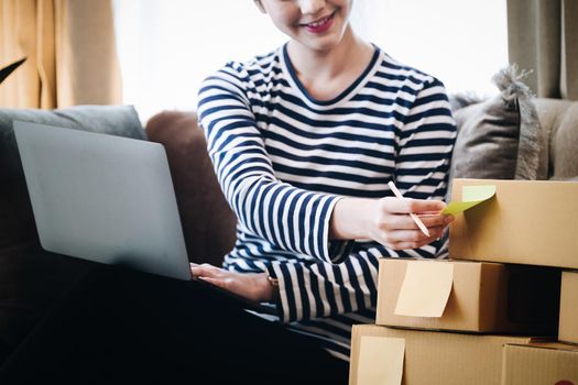 Online merchandising business idea, a beautiful girl is writing a notebook and using a computer to check the order to deliver parcels to customers according to orders received from customers