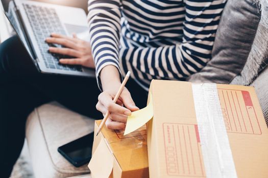 Online merchandising business idea, a beautiful girl is writing a notebook and using a computer to check the order to deliver parcels to customers according to orders received from customers