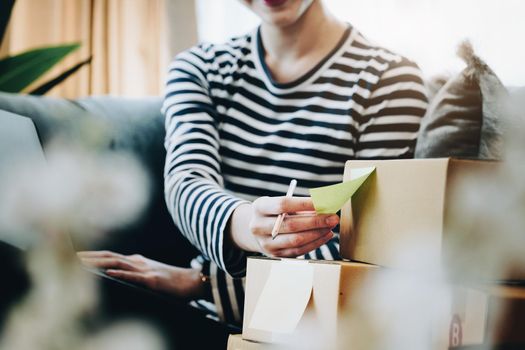 Online merchandising business idea, a beautiful girl is writing a notebook and using a computer to check the order to deliver parcels to customers according to orders received from customers