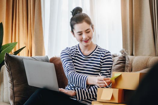 Online merchandising business idea, a beautiful girl is writing a notebook and using a computer to check the order to deliver parcels to customers according to orders received from customers