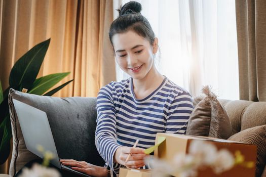 Online merchandising business idea, a beautiful girl is writing a notebook and using a computer to check the order to deliver parcels to customers according to orders received from customers